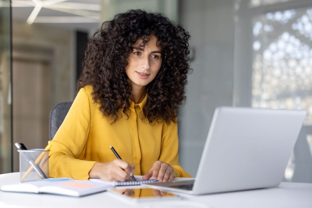 Eine Frau sitzt an einem Schreibtisch, macht Notizen und lernt online mit einem Laptop. E-Learning Anbieter bieten praxisnahe Weiterbildung für Unternehmen.